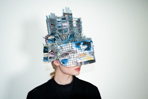 The artist wears an elaborately decorated helmet that features pieces of mirror that reflect back on the viewer
